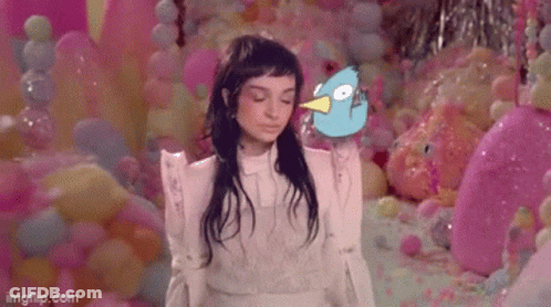 A Woman In A White Dress Is Standing In Front Of A Wall Of Pink Balloons Holding A Blue Bird . GIF