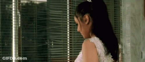 A Woman In A White Dress Is Standing In Front Of A Window With Blinds On It . GIF