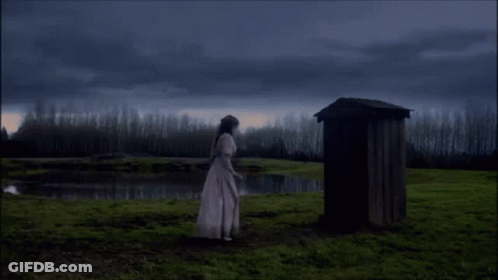 A Woman In A White Dress Is Standing In Front Of A Wooden Outhouse GIF
