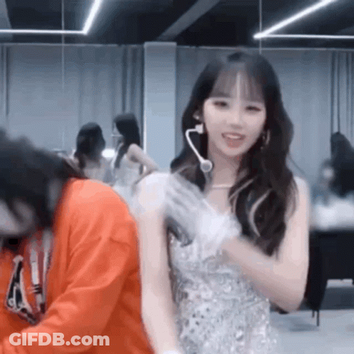 A Woman In A White Dress Is Standing Next To A Man In An Orange Sweatshirt In A Room . GIF