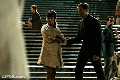 A Woman In A White Dress Is Standing Next To A Man On A Set Of Stairs GIF