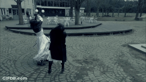 A Woman In A White Dress Is Standing On A Cobblestone Walkway GIF
