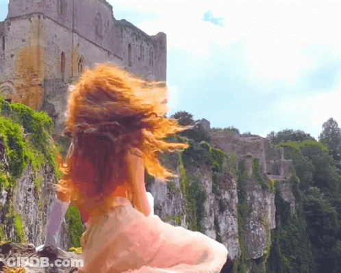 A Woman In A White Dress Is Standing On A Rocky Cliff With A Castle In The Background GIF