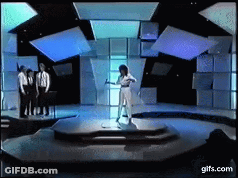 A Woman In A White Dress Is Standing On A Stage In Front Of A Group Of Men In Suits . GIF