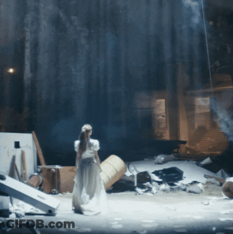 A Woman In A White Dress Is Standing On A Stage With A Barrel In The Background GIF