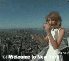 A Woman In A White Dress Is Standing On Top Of A Building With The Words Welcome To New York Below Her GIF