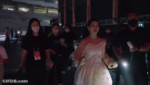 A Woman In A White Dress Is Surrounded By People Wearing Masks And A Man Holding A Flashlight .. GIF