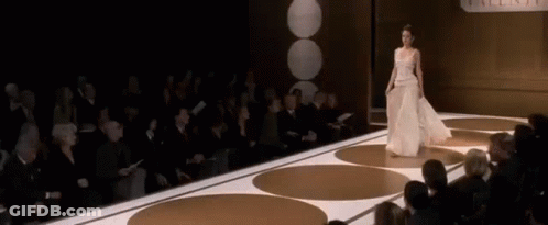 A Woman In A White Dress Is Walking Down A Runway In Front Of A Crowd Of People . GIF