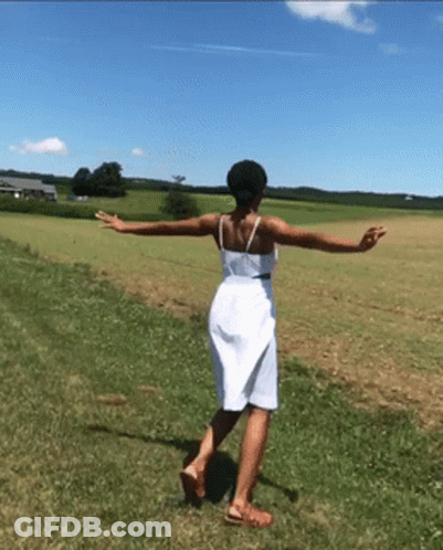 A Woman In A White Dress Is Walking Through A Grassy Field GIF