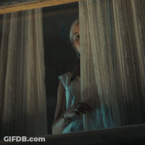 A Woman In A White Dress Looks Out A Window At Night GIF