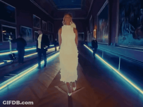 A Woman In A White Dress Walks Down A Hallway GIF