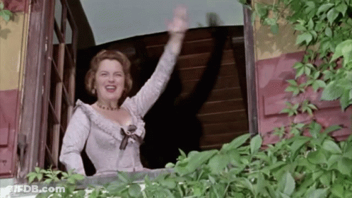 A Woman In A White Dress Waving From A Balcony GIF
