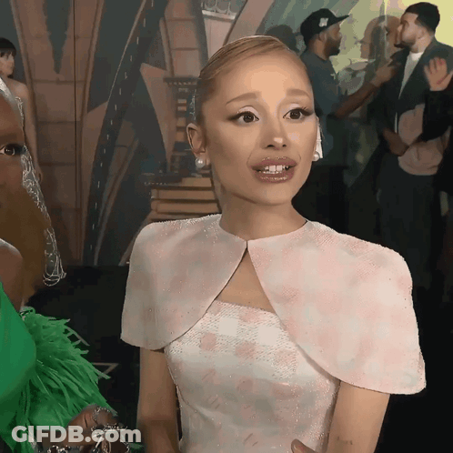 A Woman In A White Dress With A Cape On Her Shoulders Is Talking To Another Woman GIF