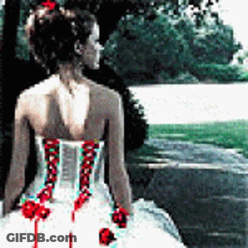 A Woman In A White Dress With Red Flowers On The Back Is Standing In Front Of A Lake GIF