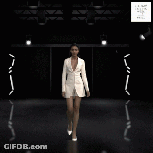 A Woman In A White Jacket And Shorts Is Walking Down A Runway At The Lakme Fashion Week GIF