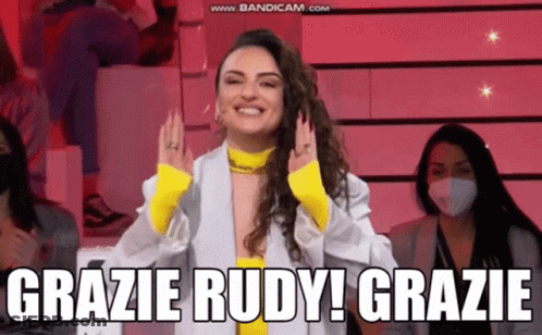A Woman In A White Jacket And Yellow Top Is Smiling And Says Grazie Rudy Grazie GIF