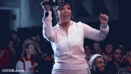 A Woman In A White Jacket Is Standing In Front Of A Crowd With Her Fist In The Air GIF
