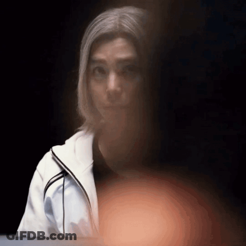 A Woman In A White Jacket Looks At The Camera With A Dark Background GIF