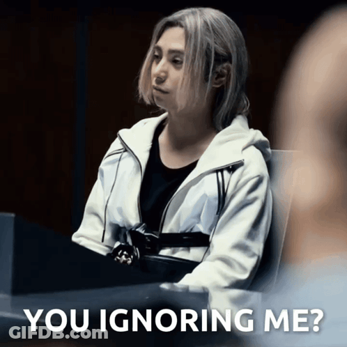 A Woman In A White Jacket Sits At A Table With The Words " You Ignoring Me " On The Bottom GIF