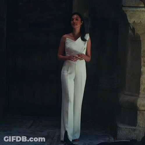 A Woman In A White Jumpsuit Is Standing In Front Of A Door GIF