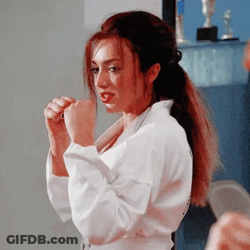 A Woman In A White Karate Uniform Is Standing In A Room With Her Hands In Her Fist . GIF