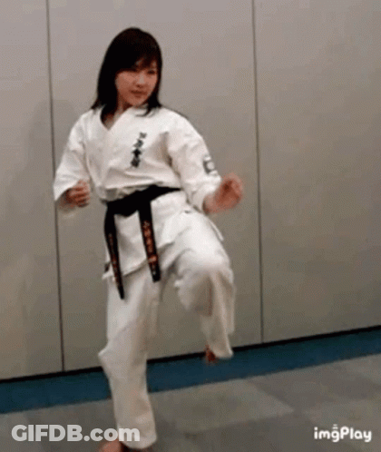 A Woman In A White Karate Uniform With A Black Belt Is Kicking A Ball . GIF