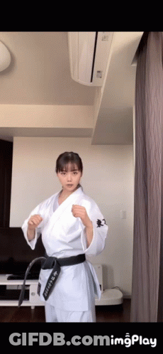 A Woman In A White Karate Uniform With A Black Belt Is Standing In A Living Room . GIF
