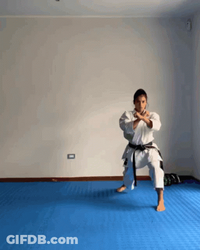 A Woman In A White Karate Uniform With A Black Belt Is Standing On A Blue Mat GIF