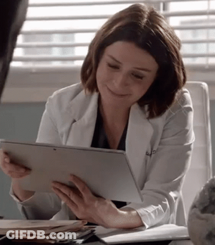 A Woman In A White Lab Coat Is Sitting At A Desk Holding A Tablet Computer . GIF