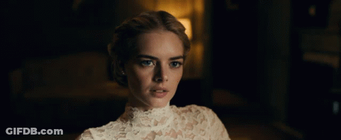A Woman In A White Lace Dress Looks At The Camera In A Dark Room GIF