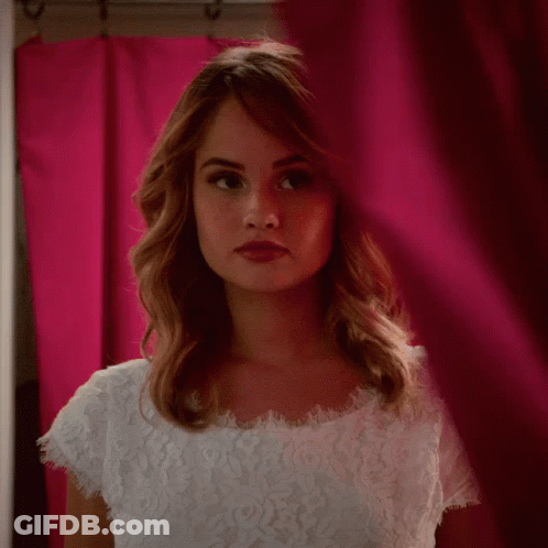 A Woman In A White Lace Top Is Looking Through A Pink Curtain GIF