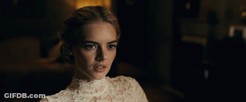A Woman In A White Lace Top Looks At The Camera In A Dark Room GIF