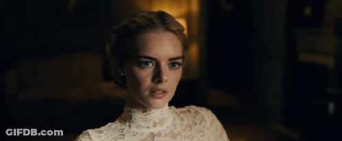 A Woman In A White Lace Top Looks At The Camera In A Dark Room GIF