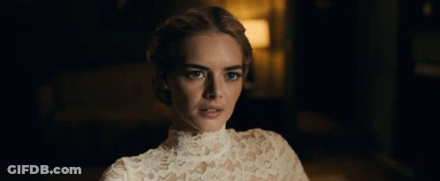 A Woman In A White Lace Top Looks At The Camera In A Dark Room GIF