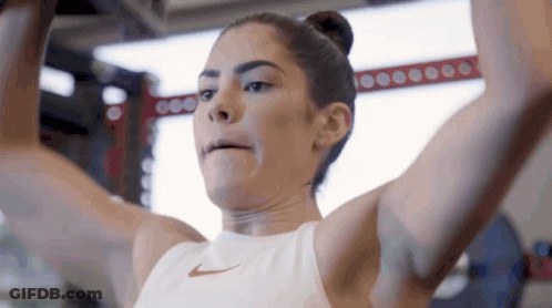 A Woman In A White Nike Tank Top Is Lifting A Barbell In A Gym GIF