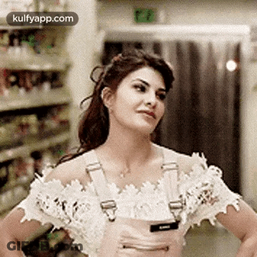 A Woman In A White Off The Shoulder Top And Pink Overalls Is Standing With Her Hands On Her Hips In A Store . GIF