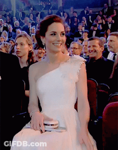 A Woman In A White One Shoulder Dress Is Smiling In Front Of A Crowd GIF