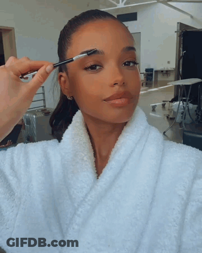 A Woman In A White Robe Has Her Eyebrows Brushed By Someone GIF