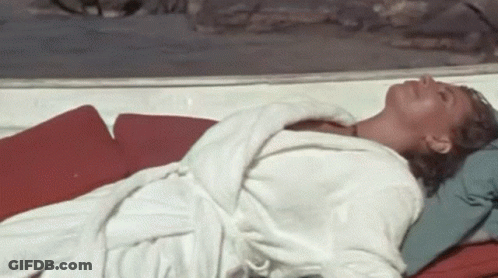 A Woman In A White Robe Is Laying On A Red Cushion On A Beach . GIF