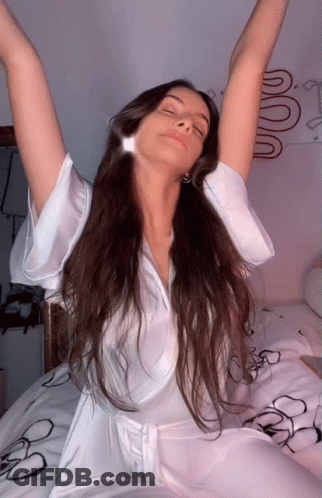 A Woman In A White Robe Is Sitting On A Bed With Her Eyes Closed GIF