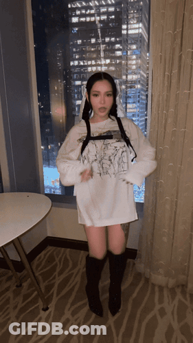 A Woman In A White Shirt And Black Boots Stands In Front Of A Window With A City In The Background GIF