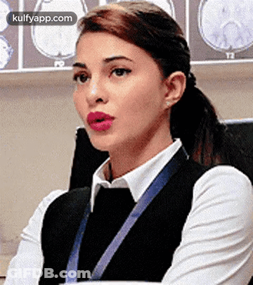 A Woman In A White Shirt And Black Vest Is Sitting In Front Of A Wall With Brain Scans . GIF