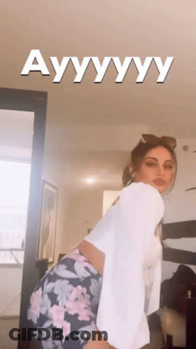 A Woman In A White Shirt And Floral Shorts Is Standing In A Room With The Words Ayyyyyy Written On The Ceiling Above Her GIF