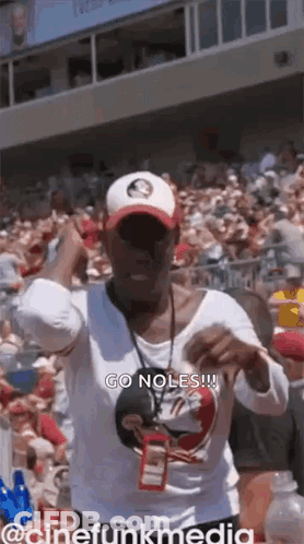 A Woman In A White Shirt And Hat Is Standing In Front Of A Crowd Of People . GIF