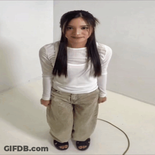 A Woman In A White Shirt And Khaki Pants Is Standing In A White Room GIF