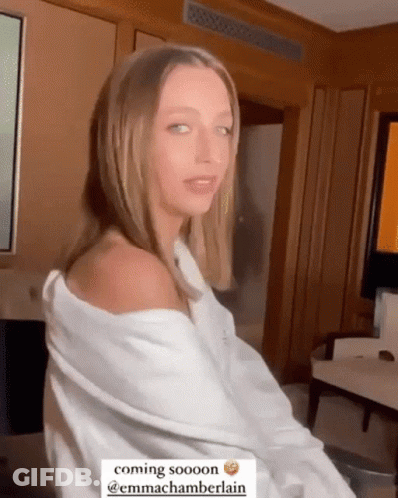 A Woman In A White Shirt Is Sitting In A Room With A Caption That Says Coming Sooooon @Emmachamberlain GIF
