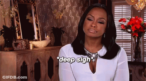 A Woman In A White Shirt Is Sitting In Front Of A Dresser And Flowers And Says Deep Sigh GIF