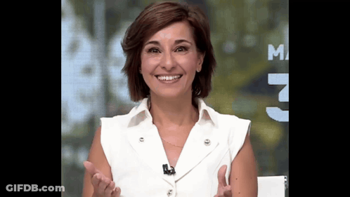 A Woman In A White Shirt Is Smiling In Front Of A Sign That Says M GIF