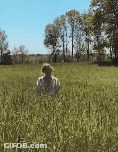 A Woman In A White Shirt Is Standing In A Grassy Field GIF