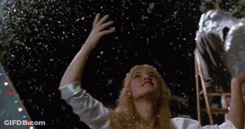 A Woman In A White Shirt Is Standing In Front Of A Christmas Tree With Snow Falling From The Sky GIF
